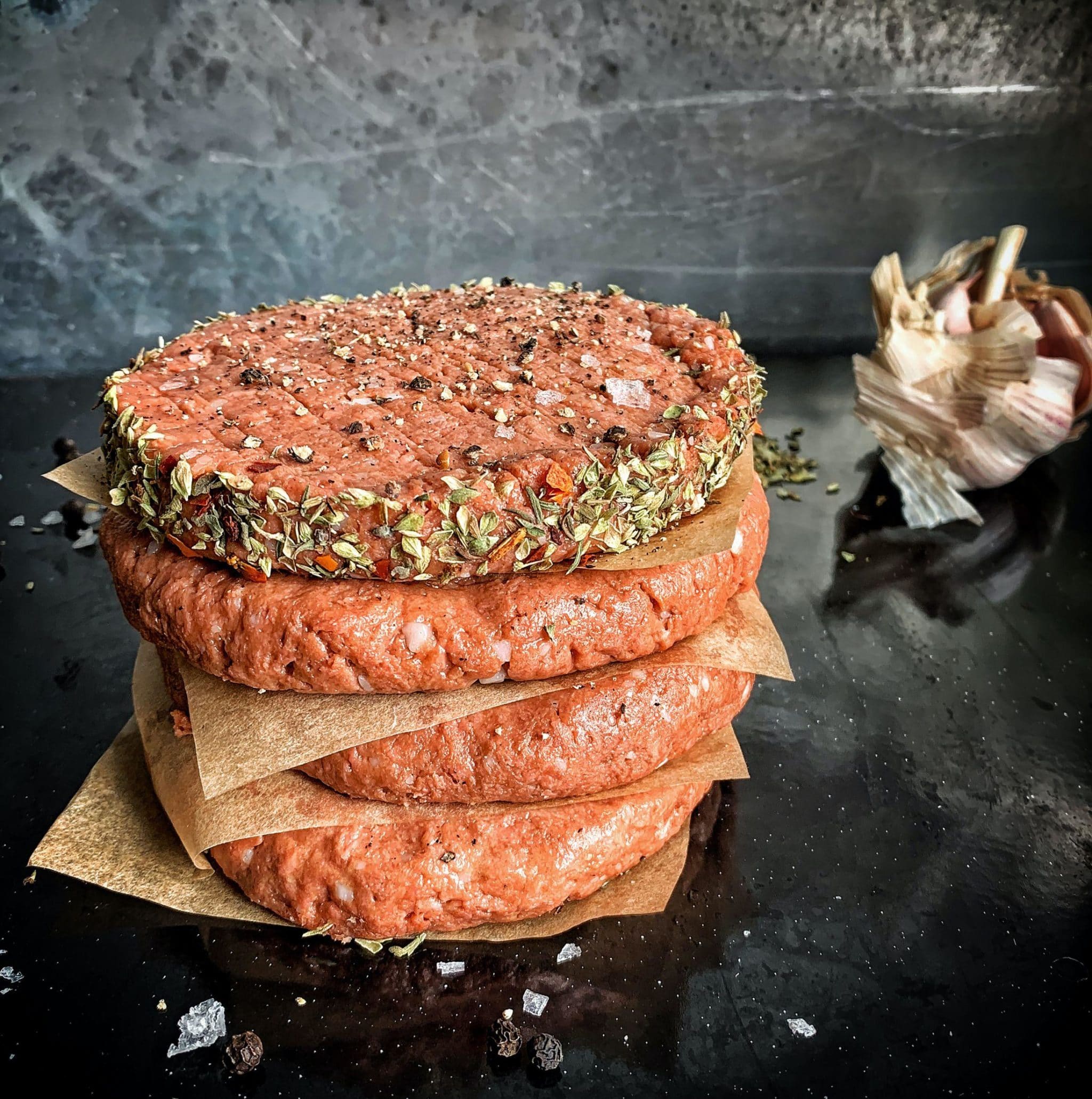 Stack of raw beef patties seasoned with herbs and spices on a black surface with garlic in the background.